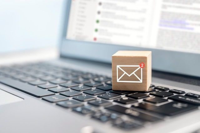 A wooden block with a white email icon and a red notification badge sitting on a laptop keyboard, with an out-of-focus email inbox on the screen.