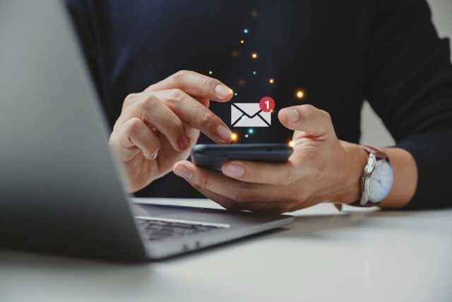 A close-up of a person's hands using a smartphone while sitting at a laptop, with a digital email icon and a red notification badge floating above the screen.
