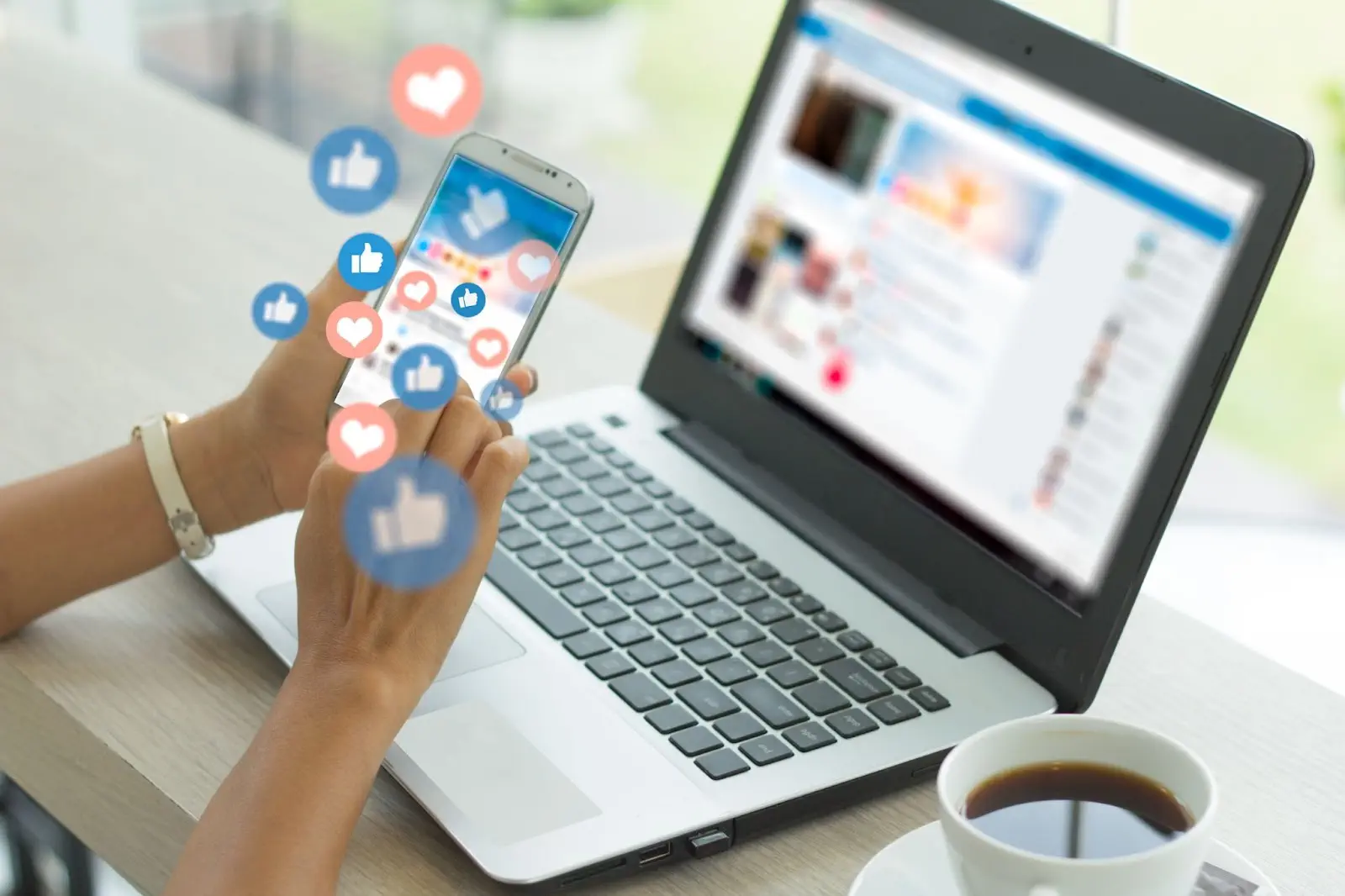 Person using a smartphone with like icons over a laptop displaying social media.