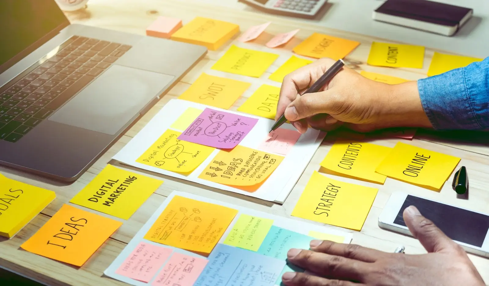 Person writing on sticky notes labeled with marketing terms, surrounded by a laptop and smartphone on a desk.