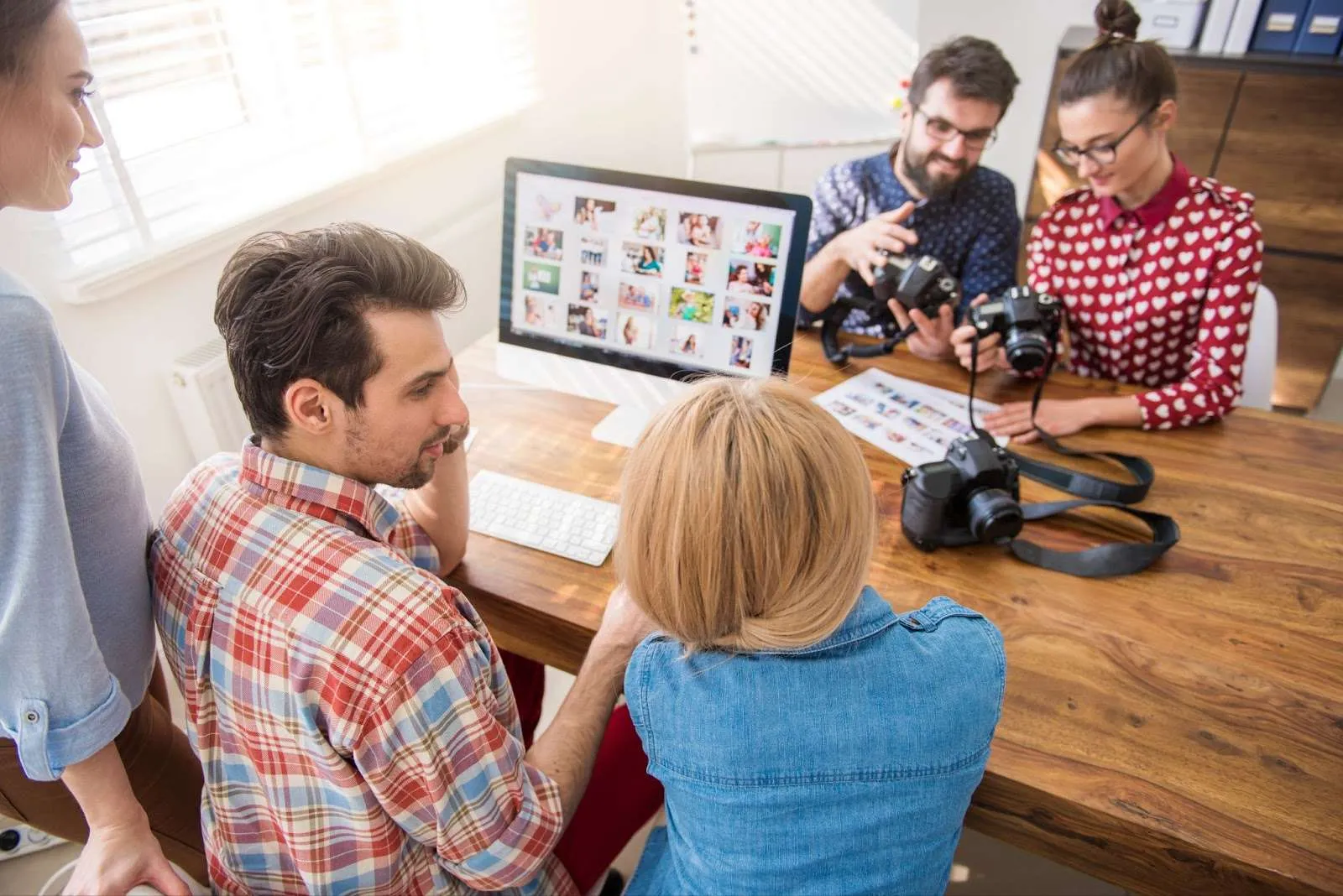 A group reviews photos on a computer and cameras at a wooden table in a bright office.