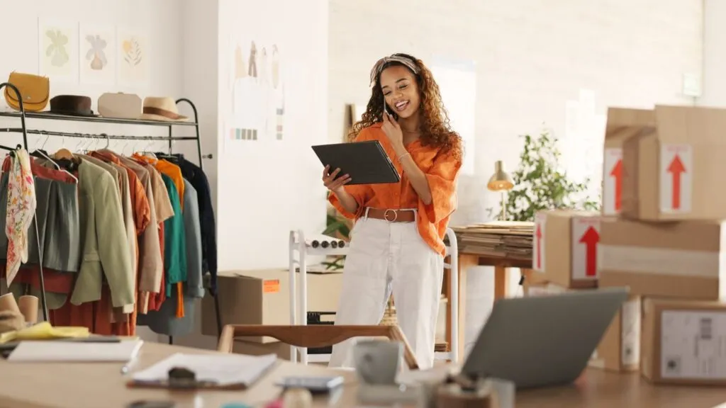 Woman in a clothing store holding a tablet and talking on the phone, surrounded by clothes and boxes.